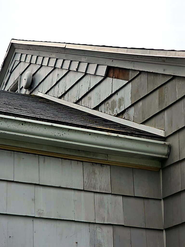 Gable-end shingles and drip edge above a low slope on a Worcester, MA cape, with missing shingle courses and exposed sheathing at the rake